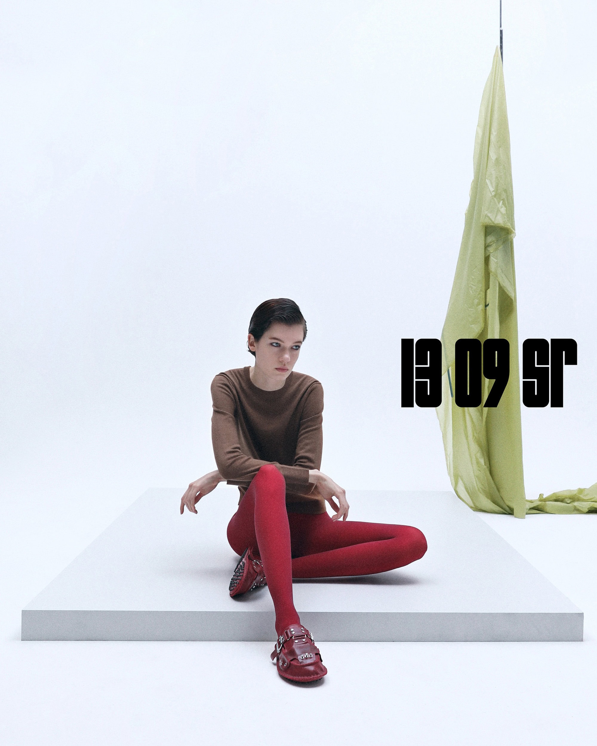 A woman with short dark hair sits on a white platform wearing a brown long-sleeve shirt, red tights, and red shoes in a bright white studio.