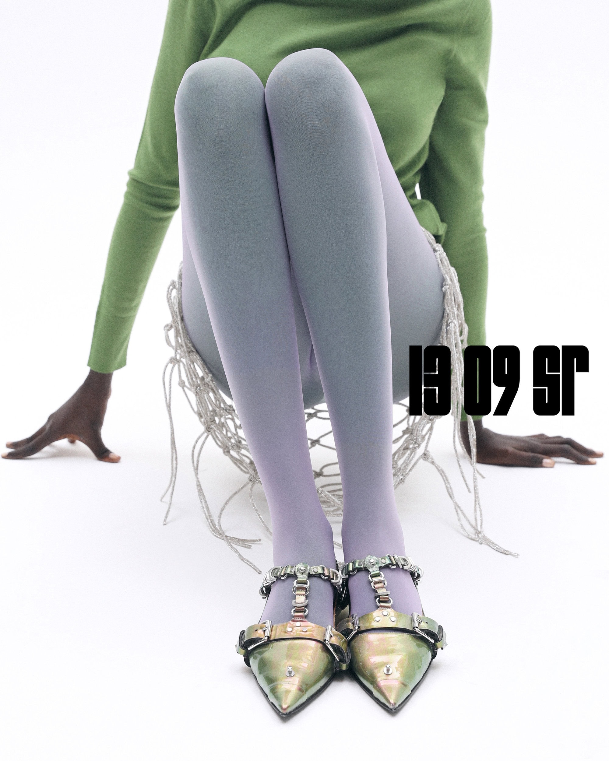 Fashion shot of a dark-skinned model wearing lilac tights and metallic gold pointed-toe heels with buckles, seated against a white backdrop.