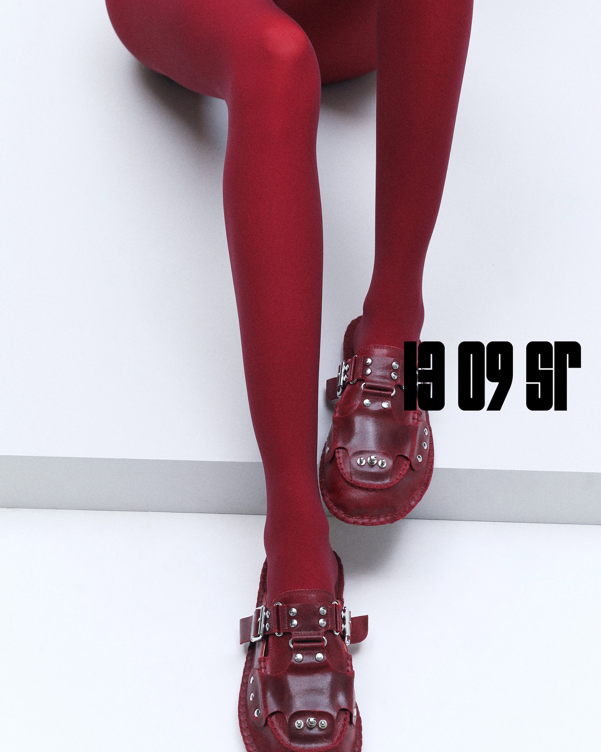A pair of red leather clogs with silver hardware, paired with red tights, are set against a clean white backdrop, highlighting the footwear.