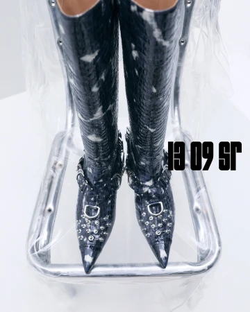 Striking black and white reptile-patterned thigh-high boots with silver studs on a reflective chrome chair against a minimalist background.