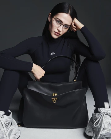 Brunette woman in black athletic wear sitting with a black leather handbag and white sneakers against a neutral gray background.