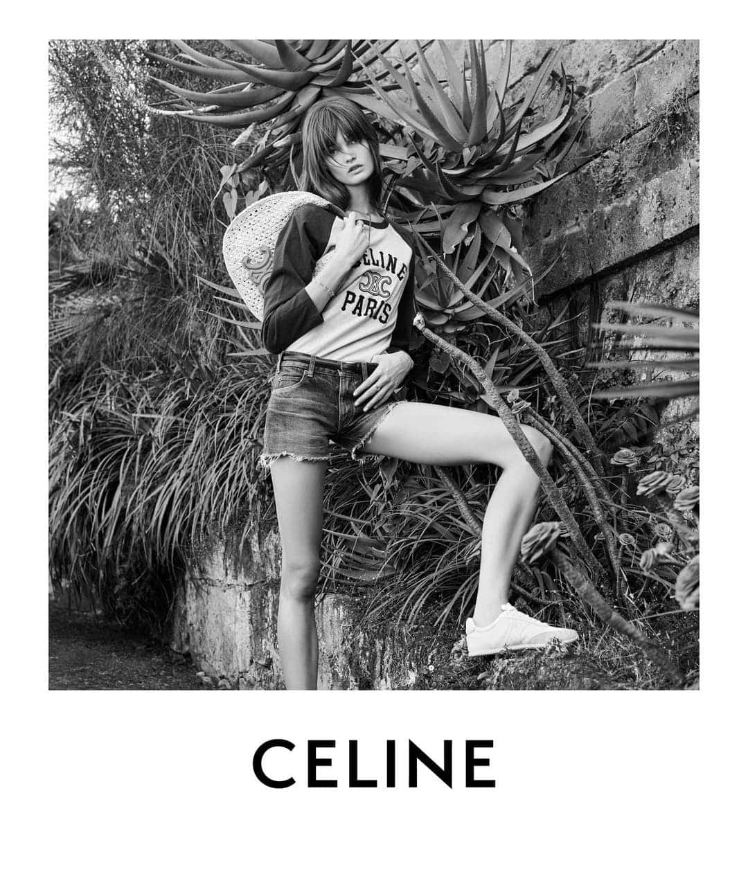 Black and white fashion shot of a woman in a logo t-shirt and jean shorts, leaning against a stone wall with leafy plants.