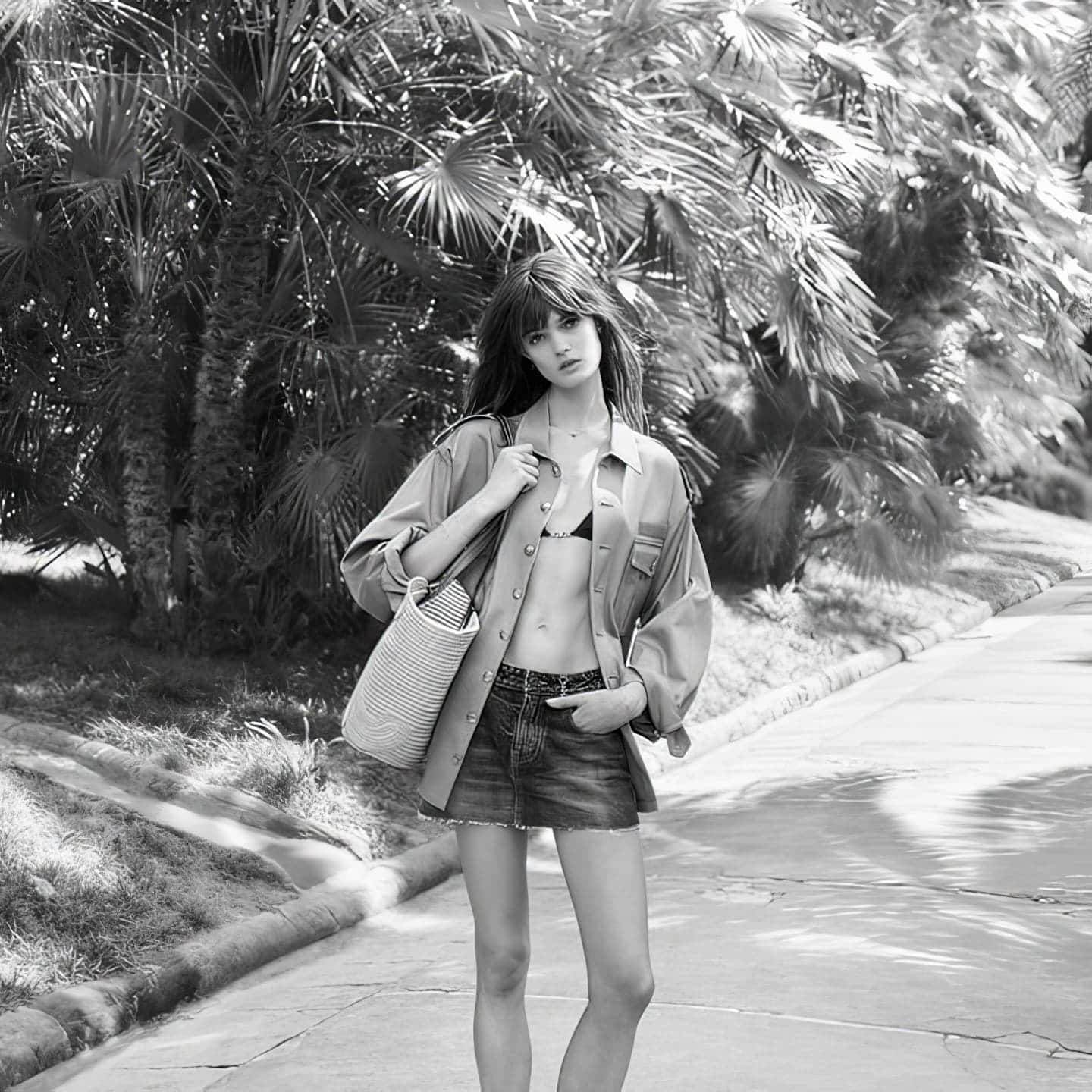 Monochrome portrait of a woman in a skirt and open shirt, strolling on a path with palm trees in the background, carrying a tote bag.