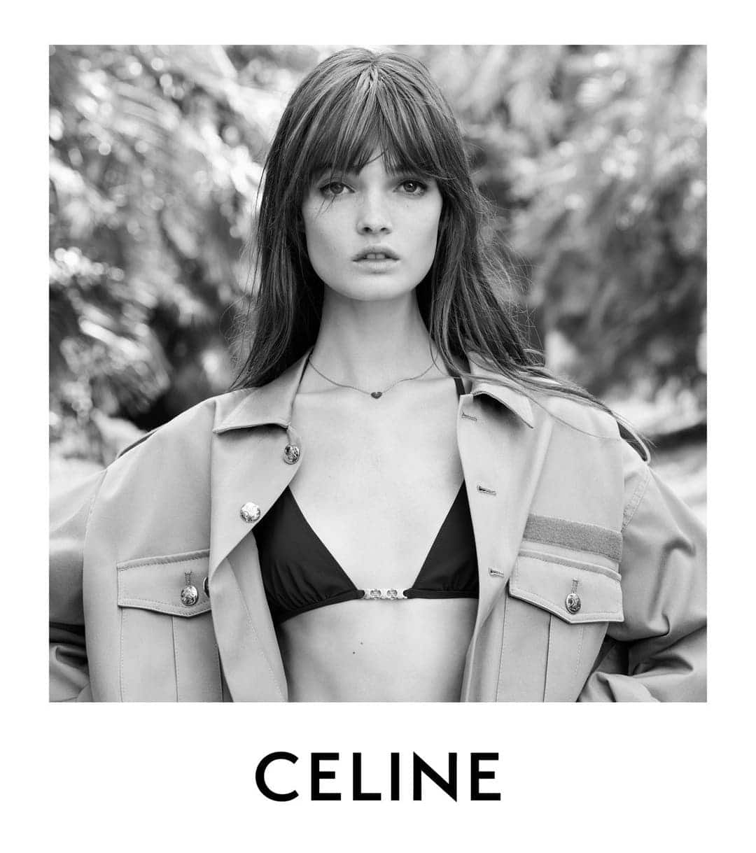 Black and white portrait of a young woman with bangs wearing a bikini top and jacket posing in front of a greenery backdrop.