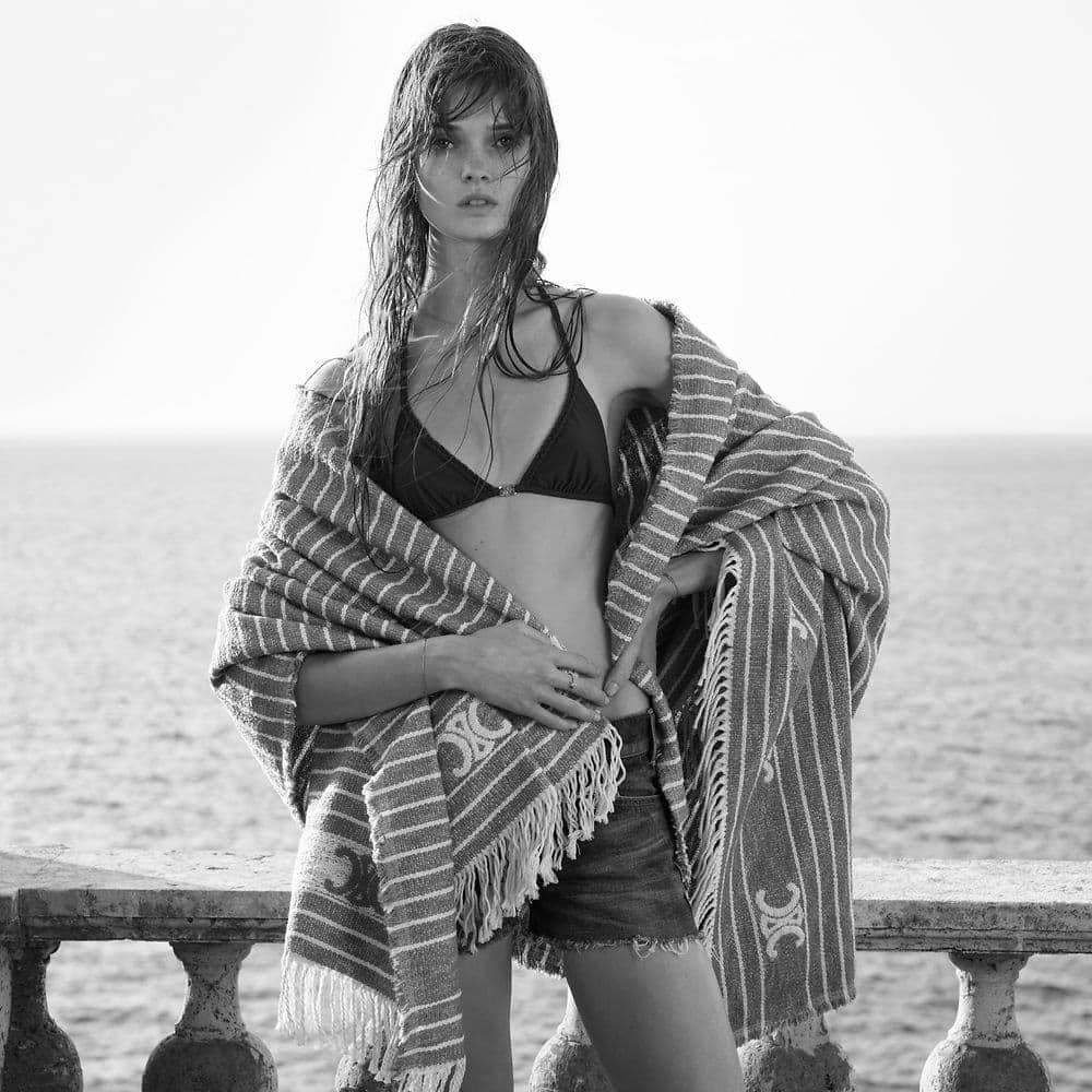 Black and white shot of an adult woman in swimwear, wrapped in a striped towel, posing on a balcony with the ocean in the background.