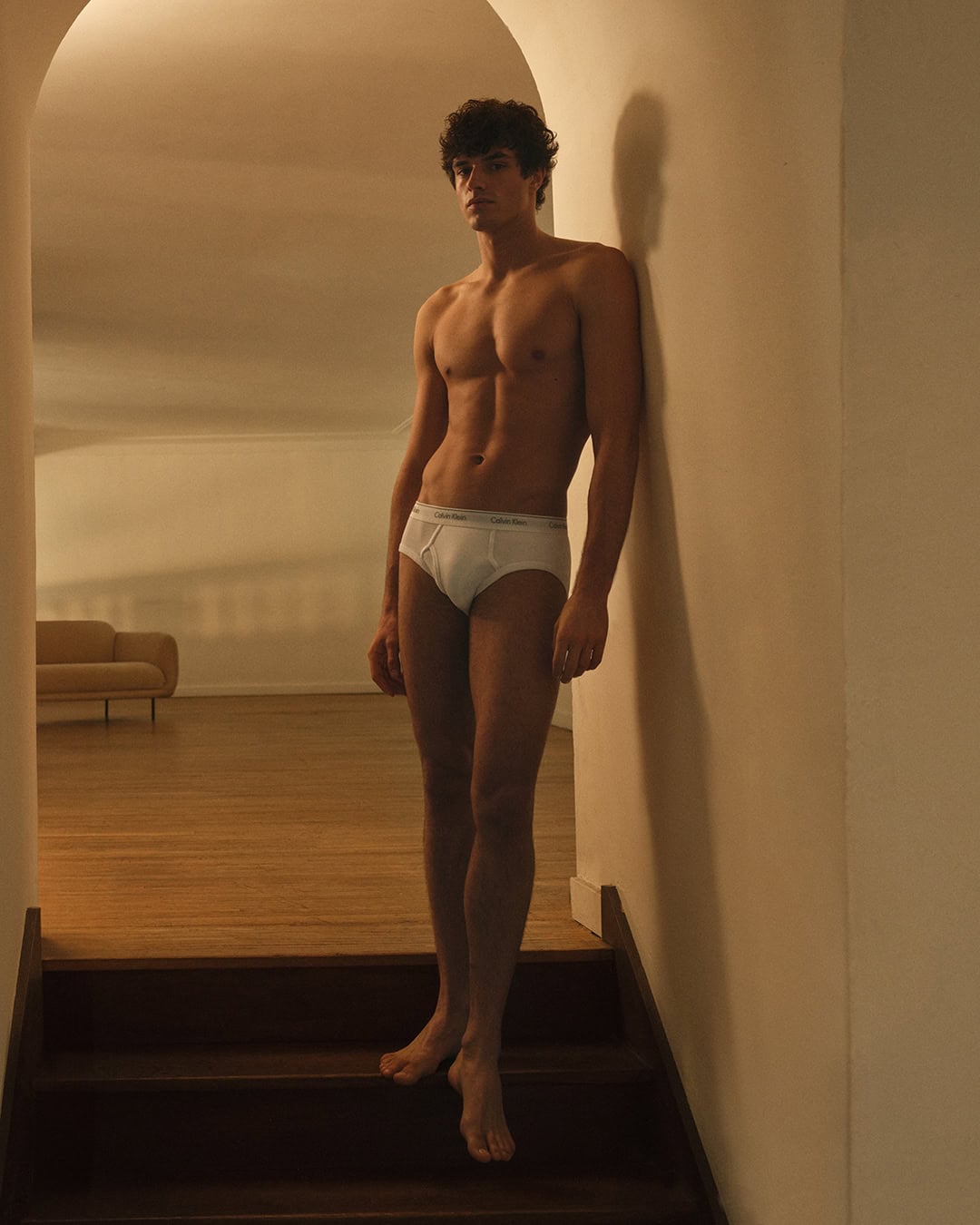Athletic man in underwear poses indoors on wooden steps against a white wall.  Warm lighting enhances the scene with a minimalist aesthetic.
