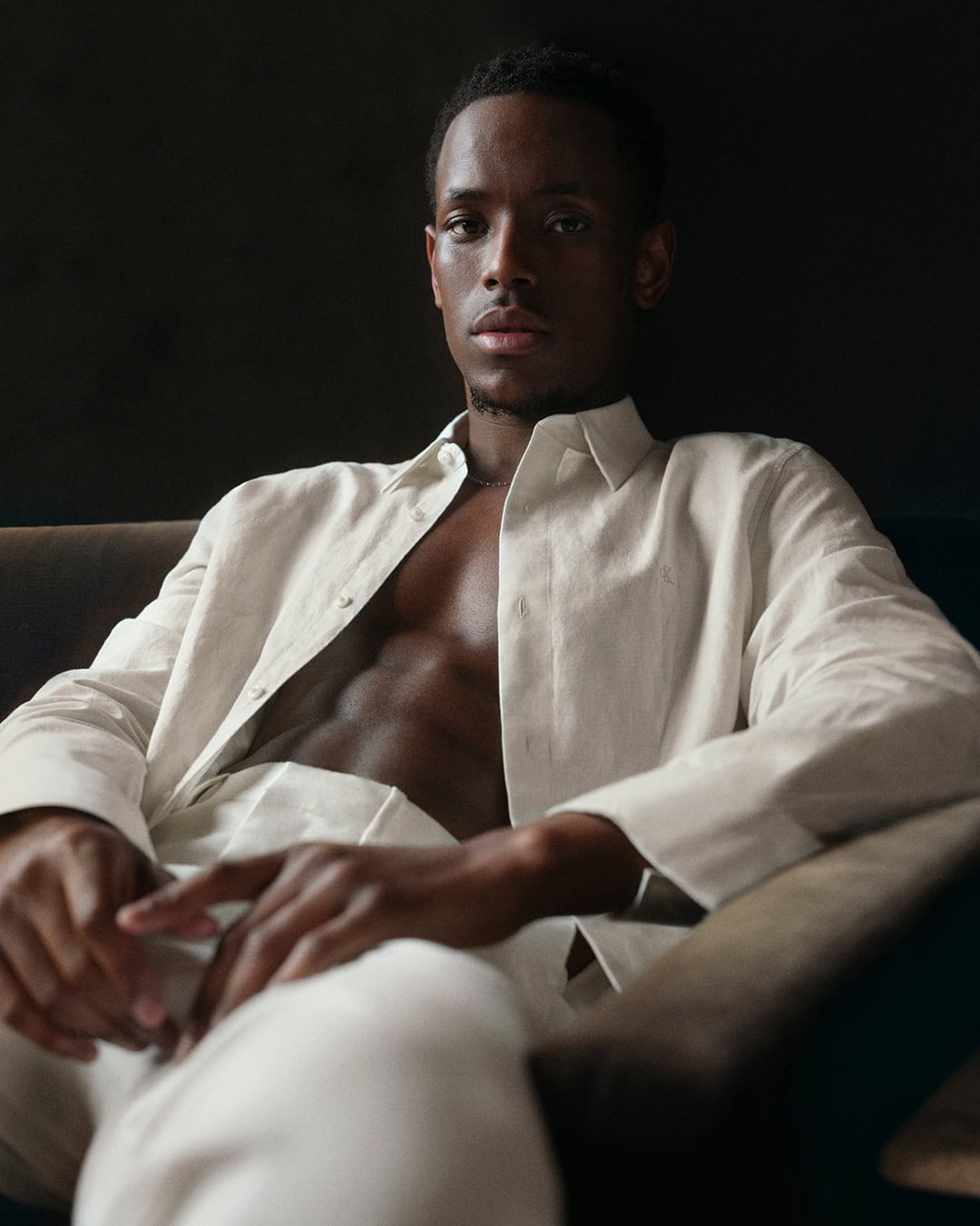 Portrait of a Black man in a relaxed pose, wearing a linen suit, indoors. The image has soft, natural lighting and a neutral color palette.