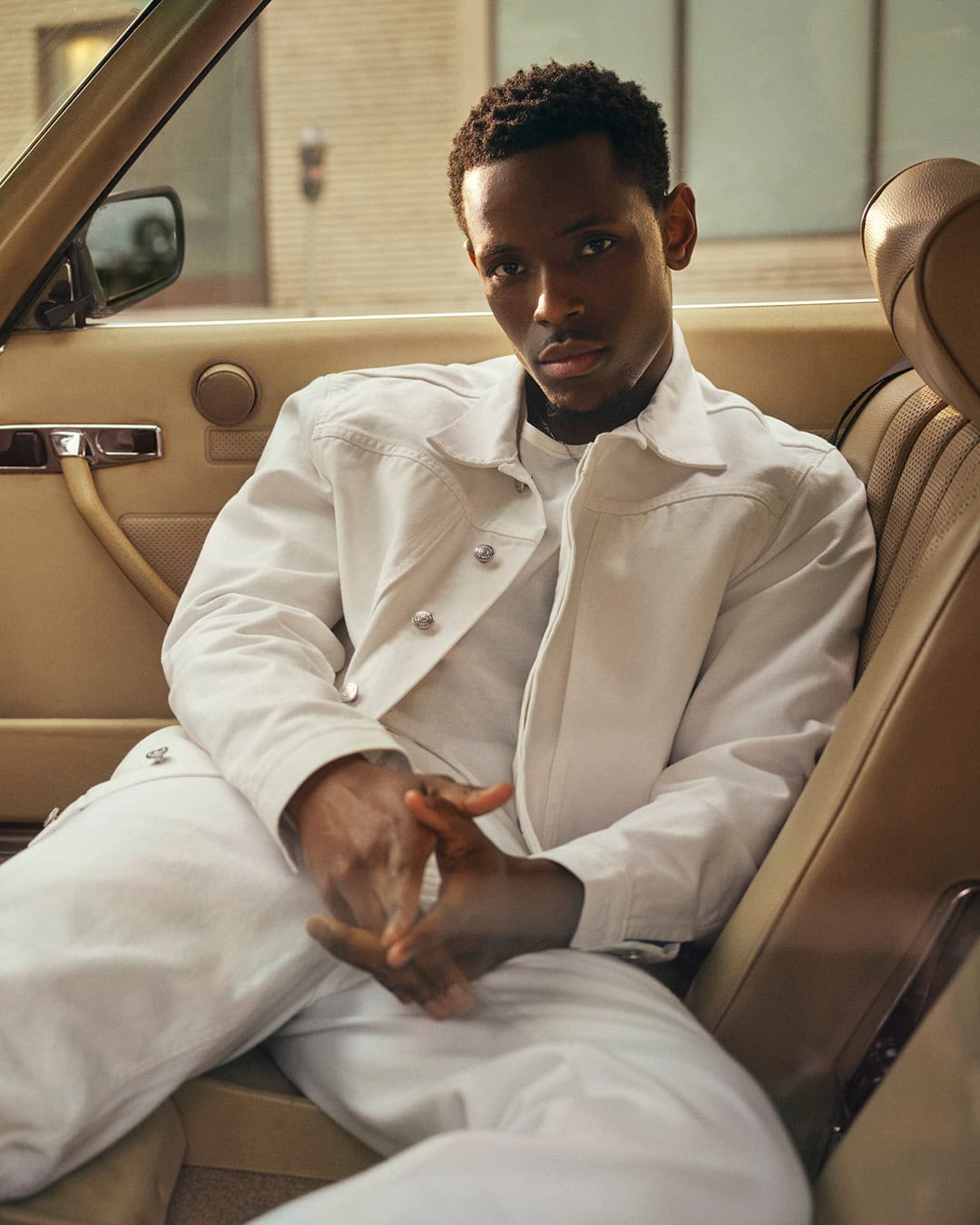 Portrait of a stylish African American man in a white denim jacket and pants posing inside a car with a beige interior.