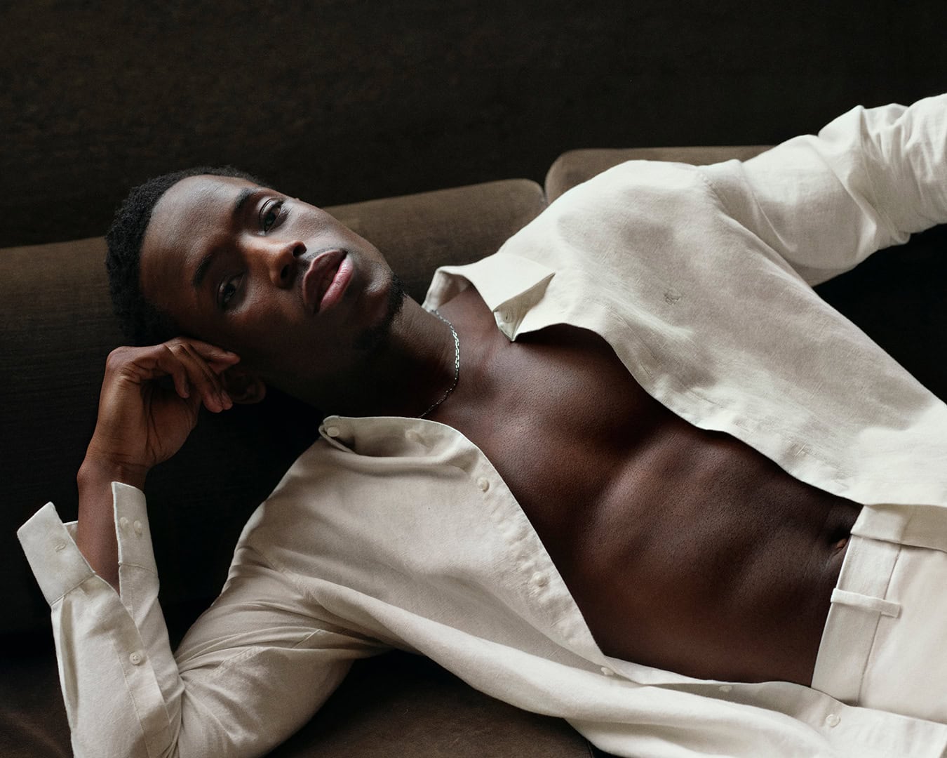 Portrait of a stylish African American man in an unbuttoned shirt and pants, casually posed on a brown sofa indoors.