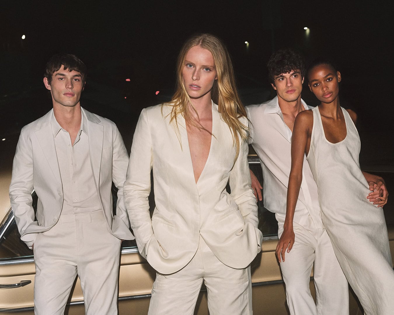 Four fashion models pose outdoors at night in cream-colored linen suits and dress, with a vintage car partially visible in the background.
