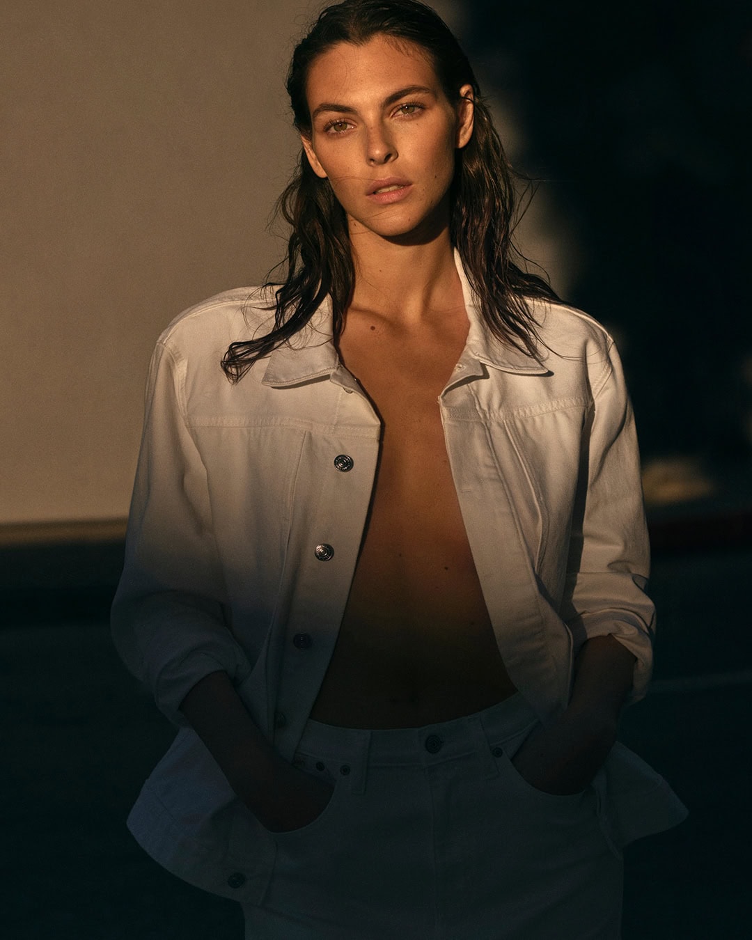 Adult female poses in a white denim jacket and jeans, hands in pockets, with sun and shadows, against a plain wall backdrop.