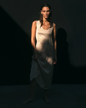 An adult woman stands in a light-filled dress against a dark background, gazing at the camera with a neutral expression and wet hair.