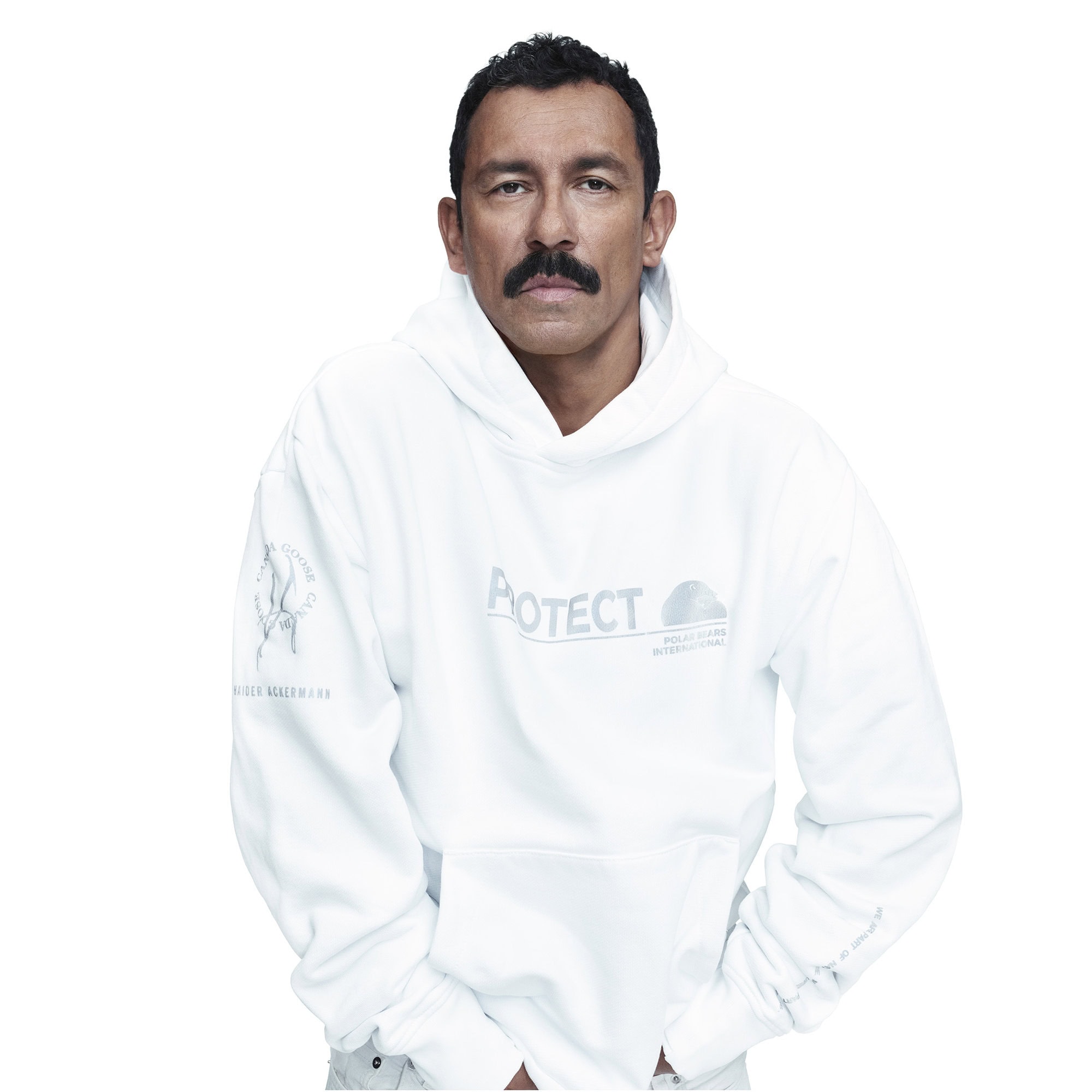 Portrait of a man with a mustache wearing a white hoodie with graphic text on a white background in a medium shot indoors.