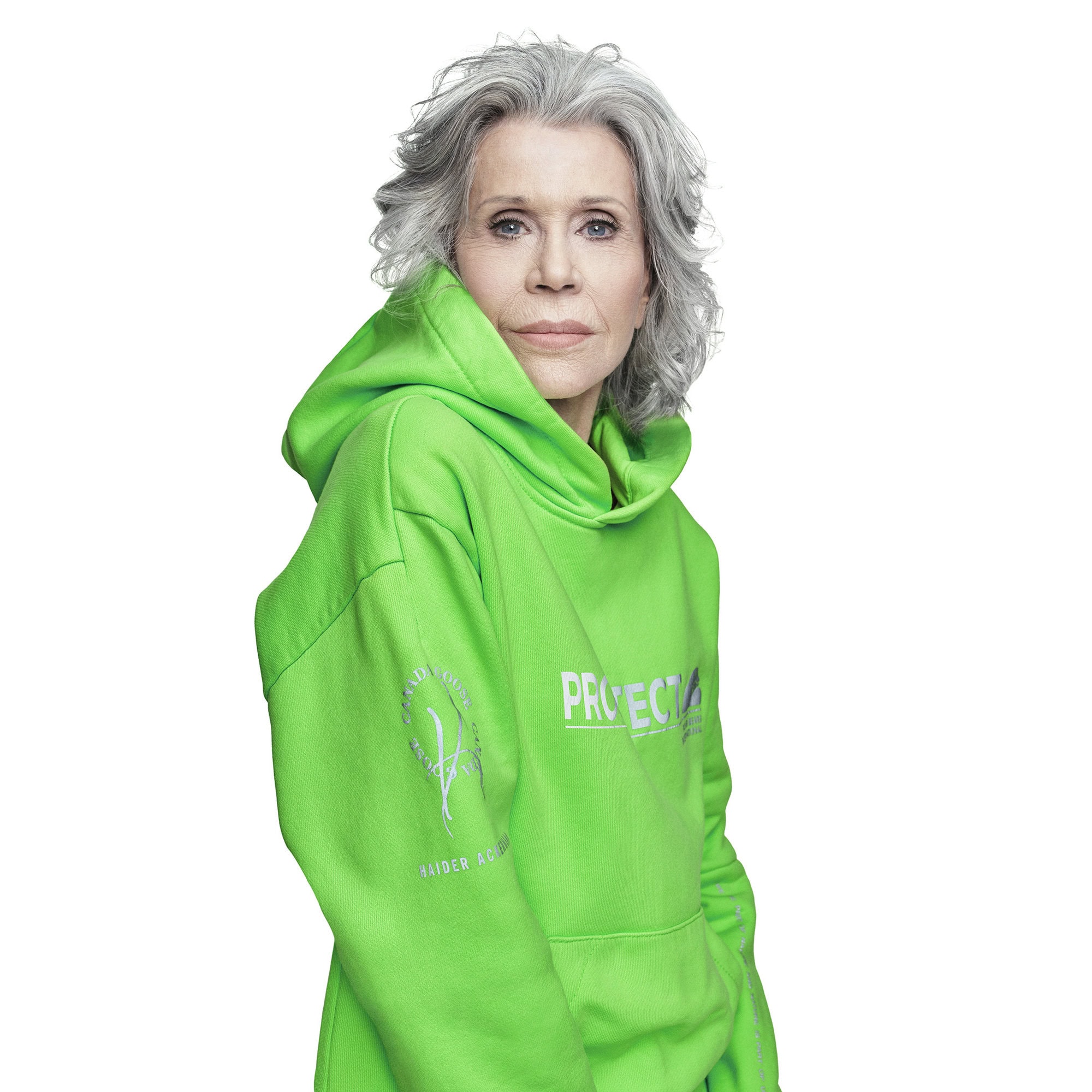 Portrait of an adult female with gray hair wearing a bright green hooded sweatshirt in a photo studio against a white backdrop.