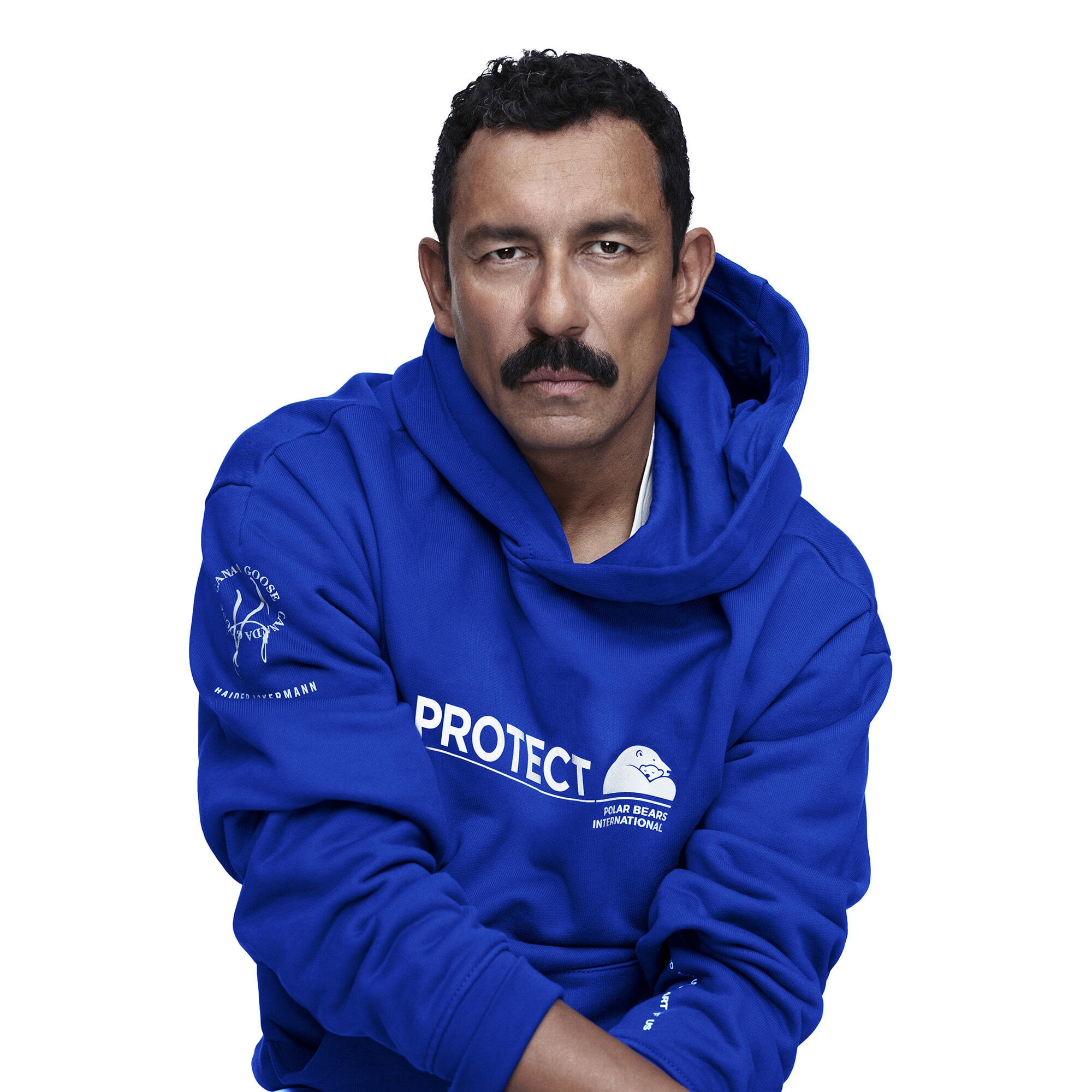 Portrait of a dark-haired man with a mustache, wearing a bright blue hoodie with the word PROTECT printed on the front, against a white backdrop.