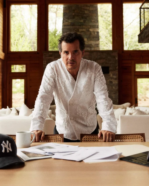 Portrait of a mature adult male leaning on a table covered with papers, wearing a patterned white jacket in a sunlit interior.
