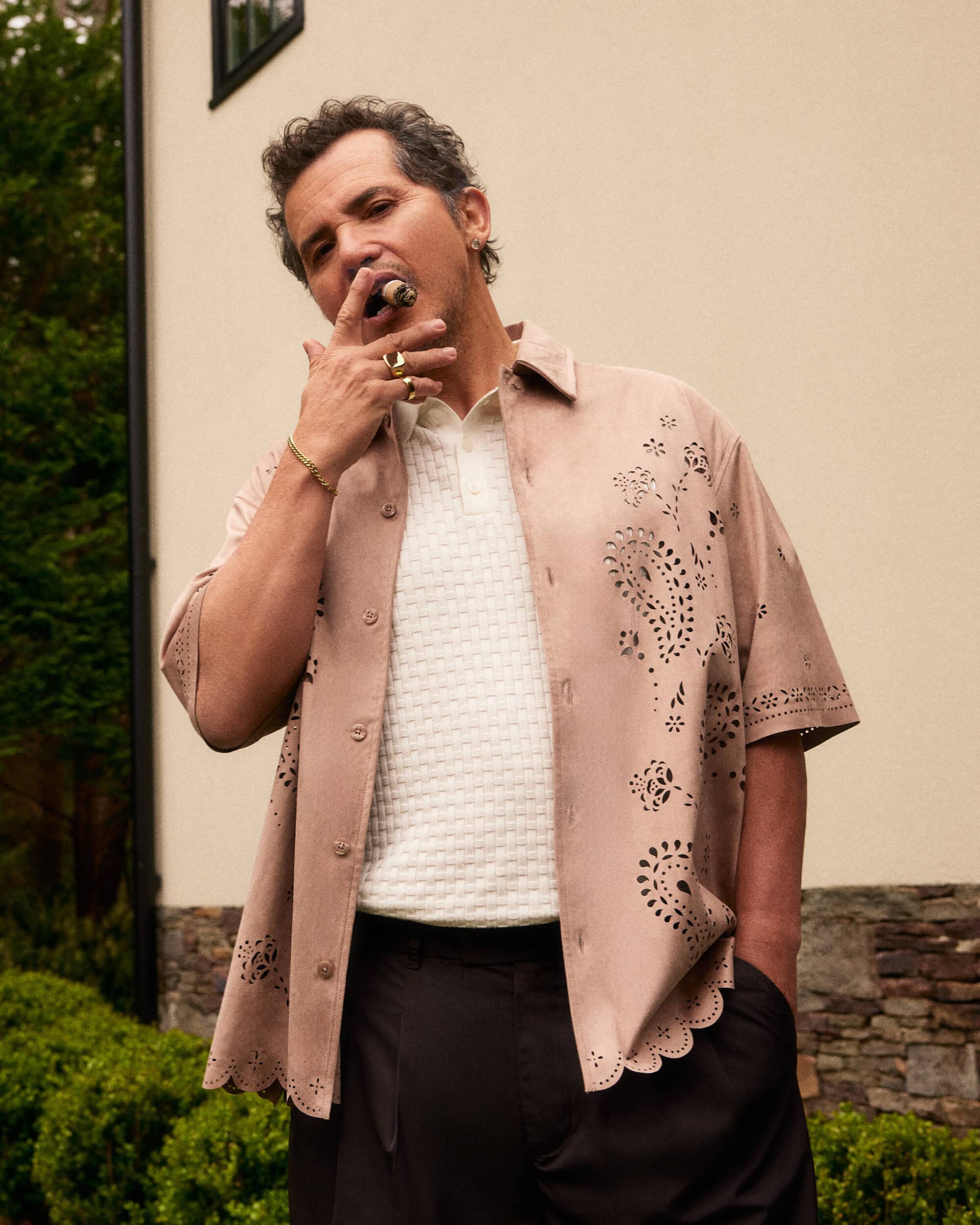 Adult male in a patterned shirt poses outside with a cigar in his mouth, wearing a white polo shirt, black slacks, and gold jewelry.