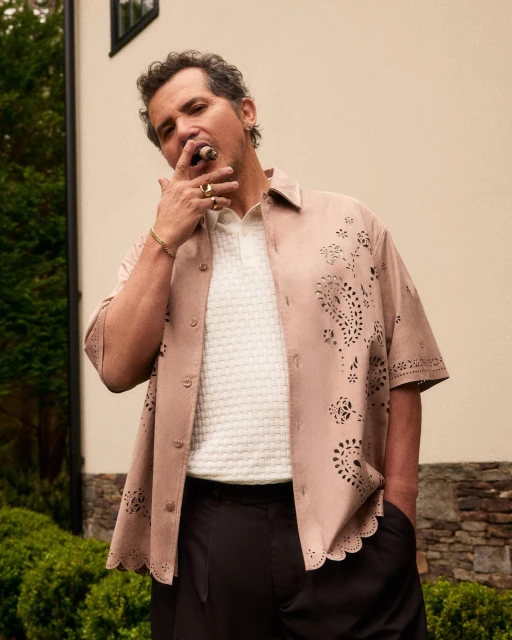 Adult male in a patterned shirt poses outside with a cigar in his mouth, wearing a white polo shirt, black slacks, and gold jewelry.