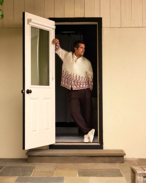 A stylish man stands in a doorway wearing a cream colored shirt with dark brown pants and white shoes, in a home setting.