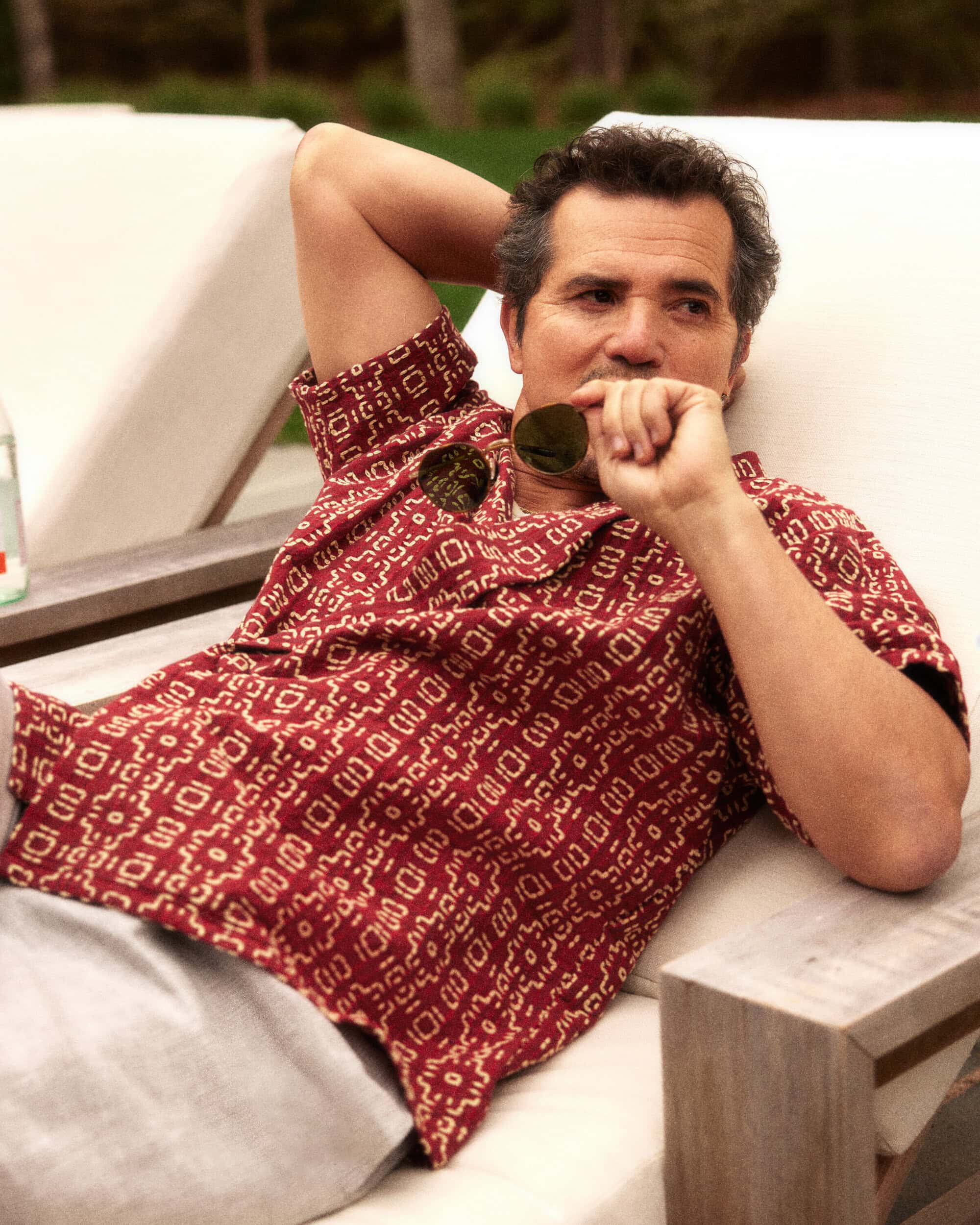 Adult male in patterned shirt, lounging outdoors with sunglasses, white daybed, and greenery in the background. Leisure lifestyle.