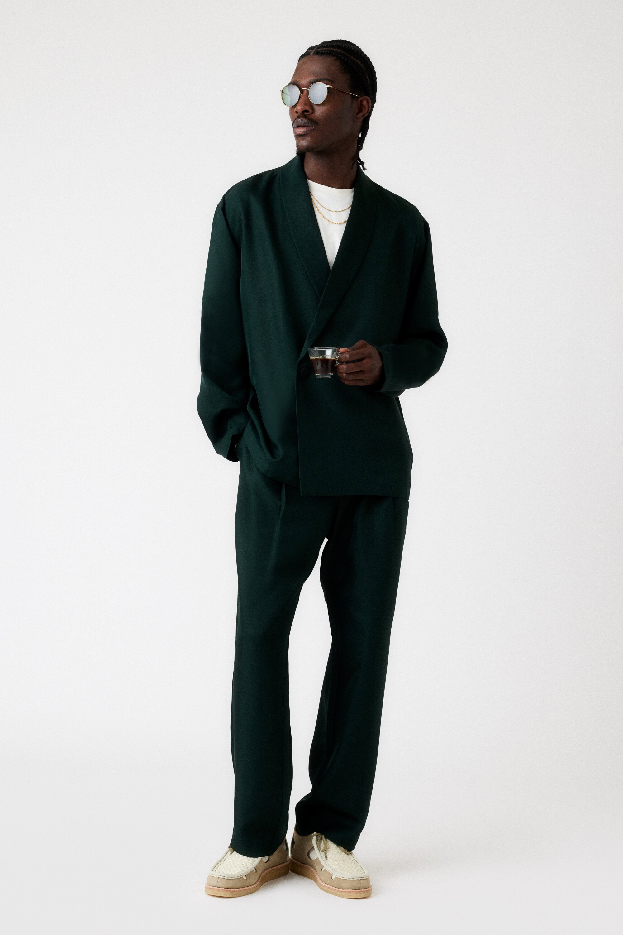 Full body shot of a stylish Black man wearing a dark green linen suit, sunglasses, gold chain, holding a vintage camera indoors.