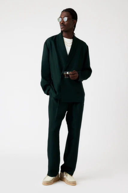 Full body shot of a stylish Black man wearing a dark green linen suit, sunglasses, gold chain, holding a vintage camera indoors.