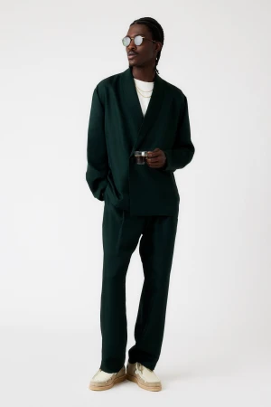 Full body shot of a stylish Black man wearing a dark green linen suit, sunglasses, gold chain, holding a vintage camera indoors.