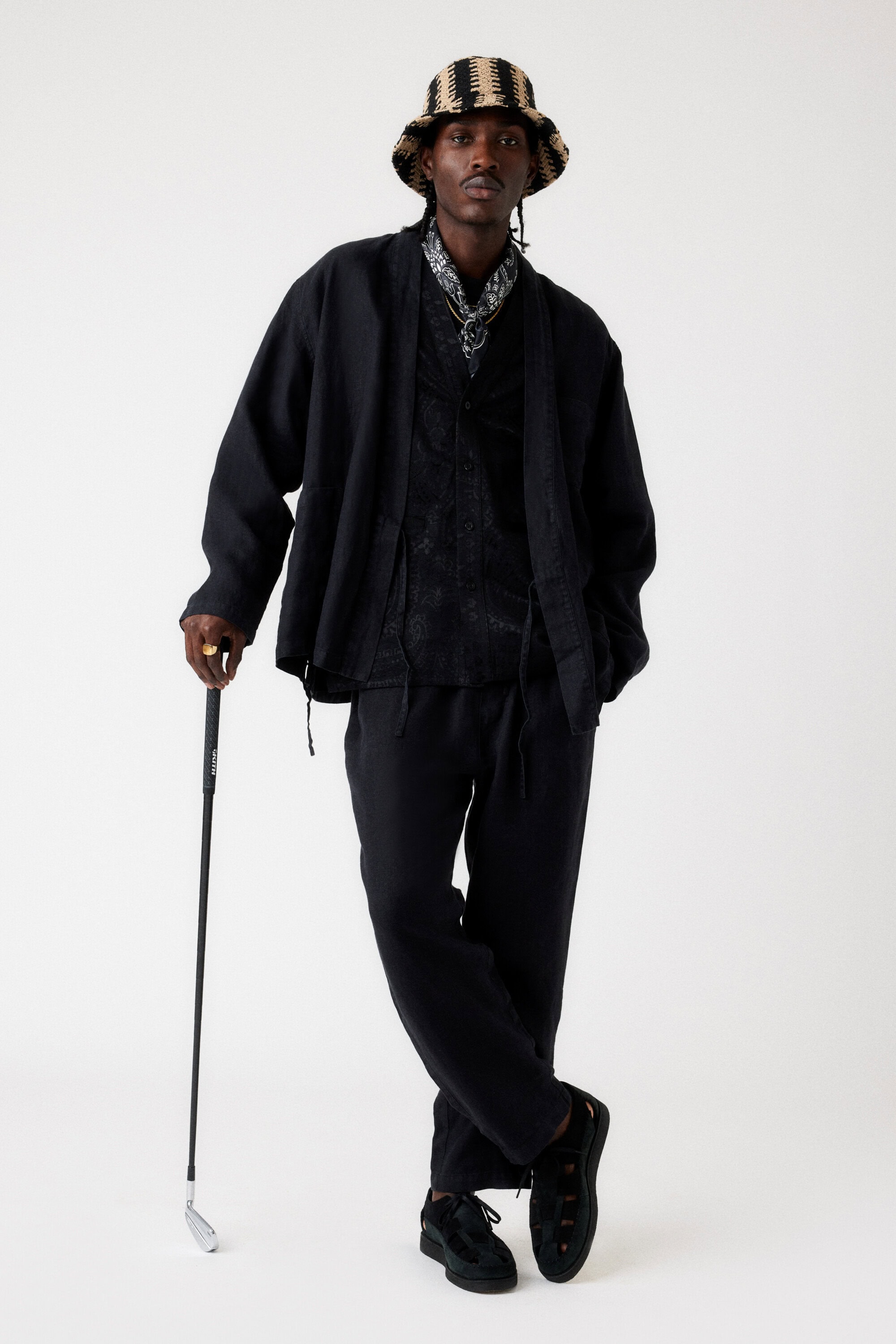 Adult Black male in stylish black suit and striped bucket hat stands with a golf club against a plain background in a studio shot.
