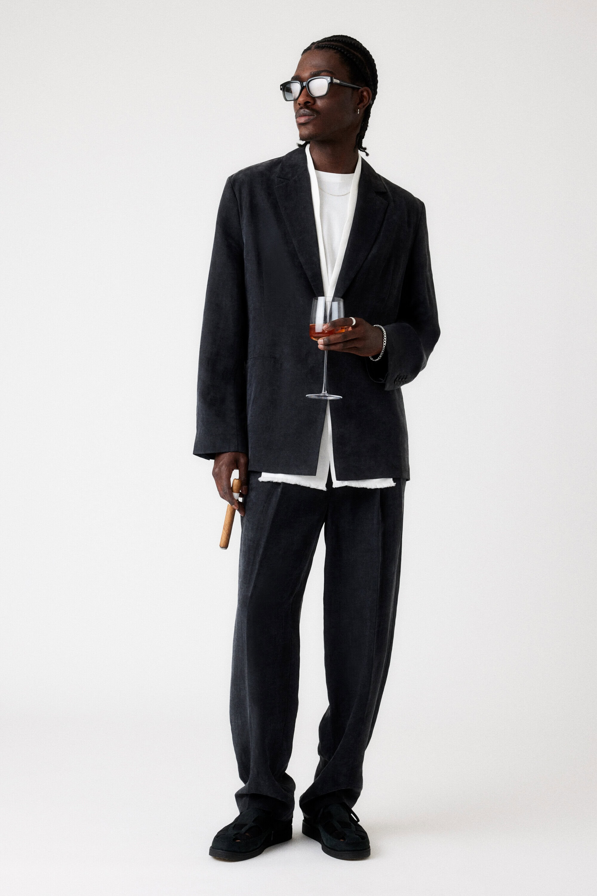 Full shot of Black male model in an oversized corduroy suit and dark glasses, holding a wine glass and cigar on a white background.