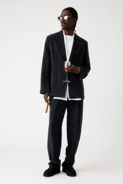 Full shot of Black male model in an oversized corduroy suit and dark glasses, holding a wine glass and cigar on a white background.