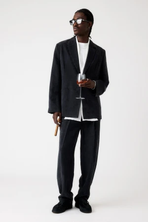 Full shot of Black male model in an oversized corduroy suit and dark glasses, holding a wine glass and cigar on a white background.