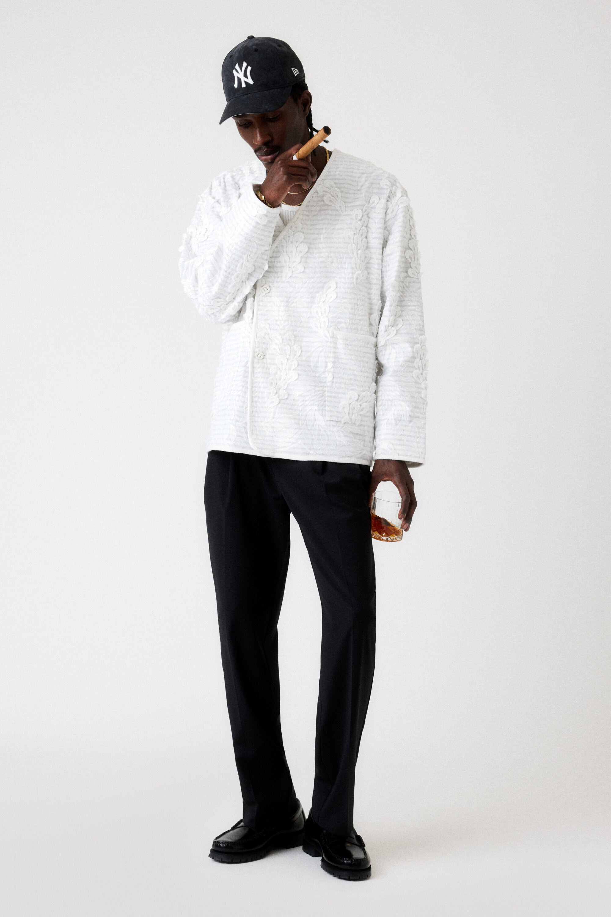 Stylish African-American man in a baseball cap, wearing a white patterned shirt and black pants, smoking a cigar and holding a glass.