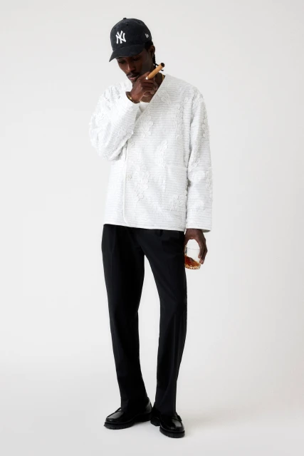 Stylish African-American man in a baseball cap, wearing a white patterned shirt and black pants, smoking a cigar and holding a glass.