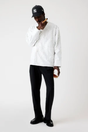 Stylish African-American man in a baseball cap, wearing a white patterned shirt and black pants, smoking a cigar and holding a glass.