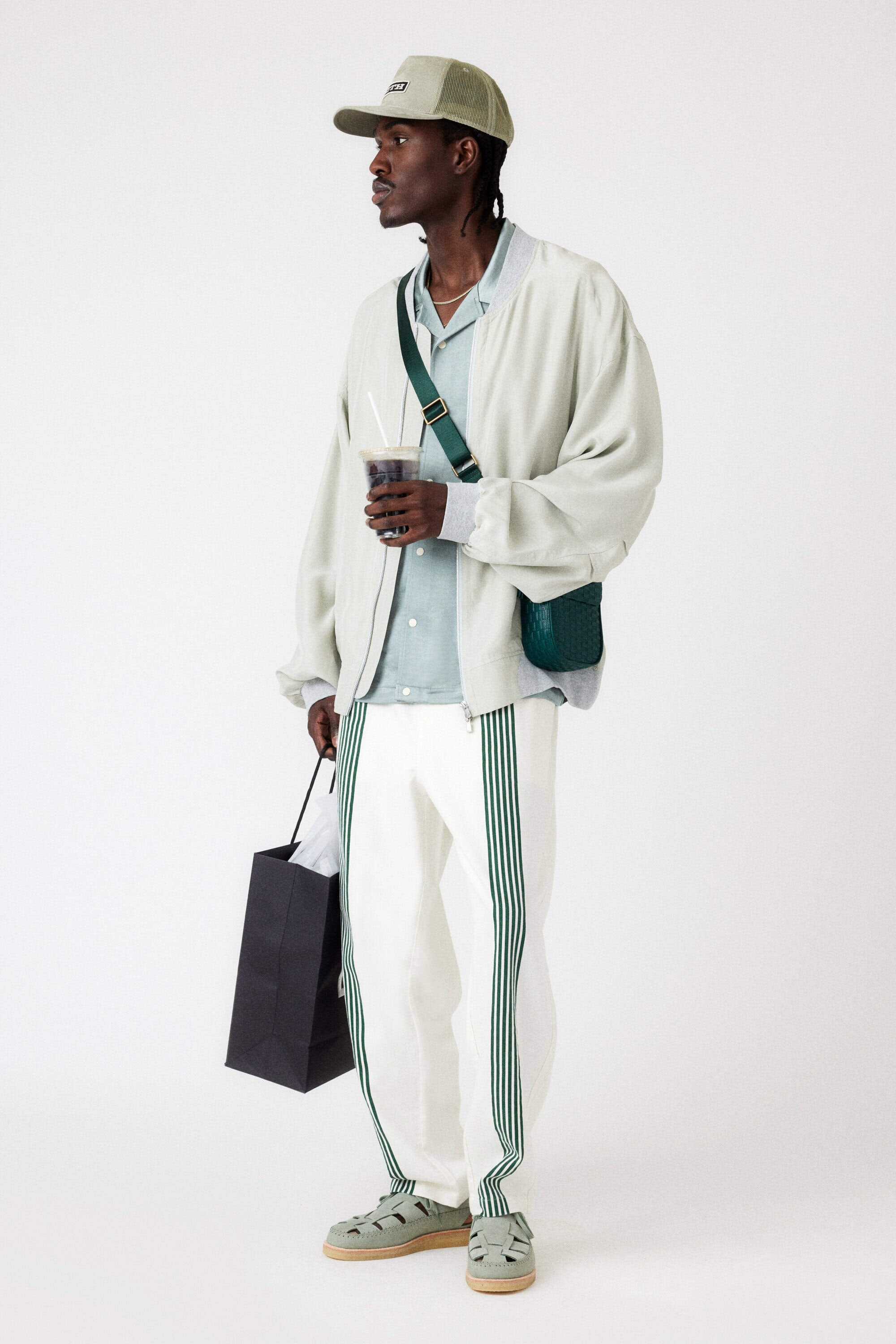 A dark-skinned male model stands in a studio wearing a light jacket, striped pants, cap, sandals, and a shoulder bag, holding a drink and bag.