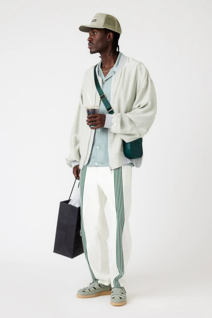 A dark-skinned male model stands in a studio wearing a light jacket, striped pants, cap, sandals, and a shoulder bag, holding a drink and bag.