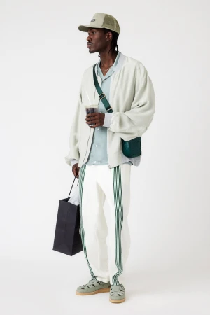 A dark-skinned male model stands in a studio wearing a light jacket, striped pants, cap, sandals, and a shoulder bag, holding a drink and bag.