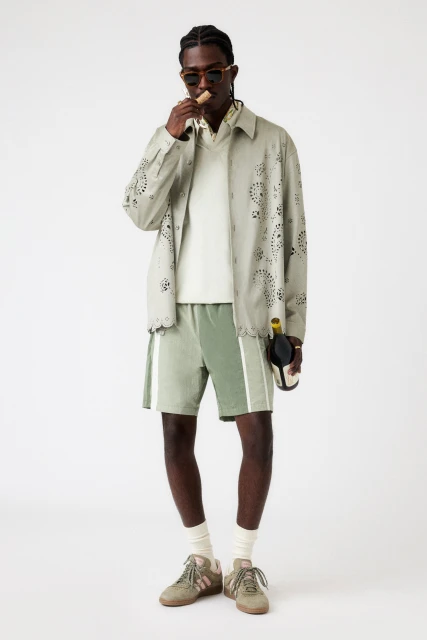 Stylish man in light green patterned jacket and shorts holding wine bottle. Fashion editorial shot against white background.