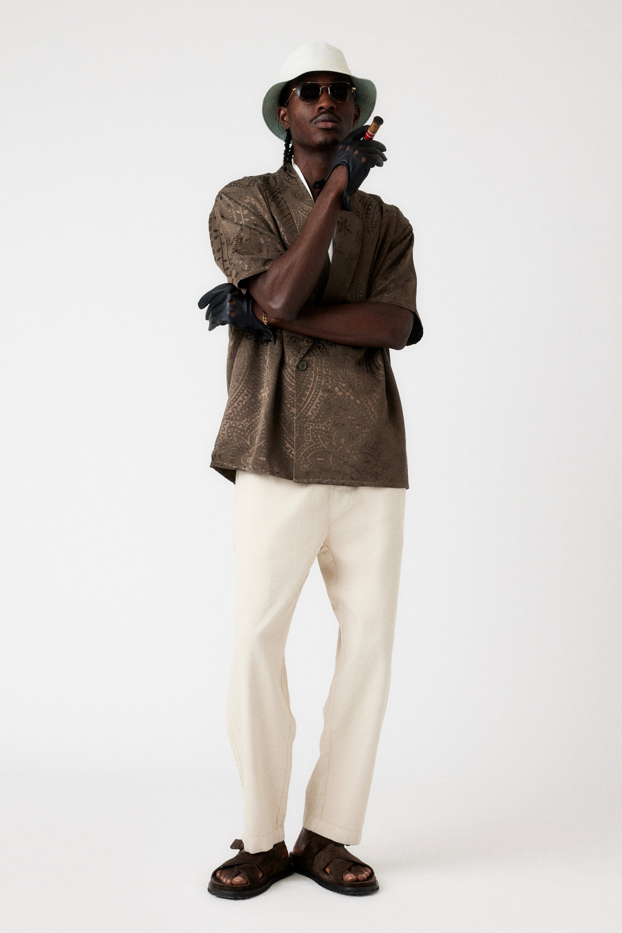 Full shot of a Black adult male in a brown patterned shirt, beige pants, sunglasses, hat and leather gloves smoking a cigar.