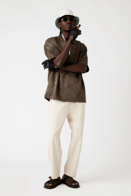 Full shot of a Black adult male in a brown patterned shirt, beige pants, sunglasses, hat and leather gloves smoking a cigar.