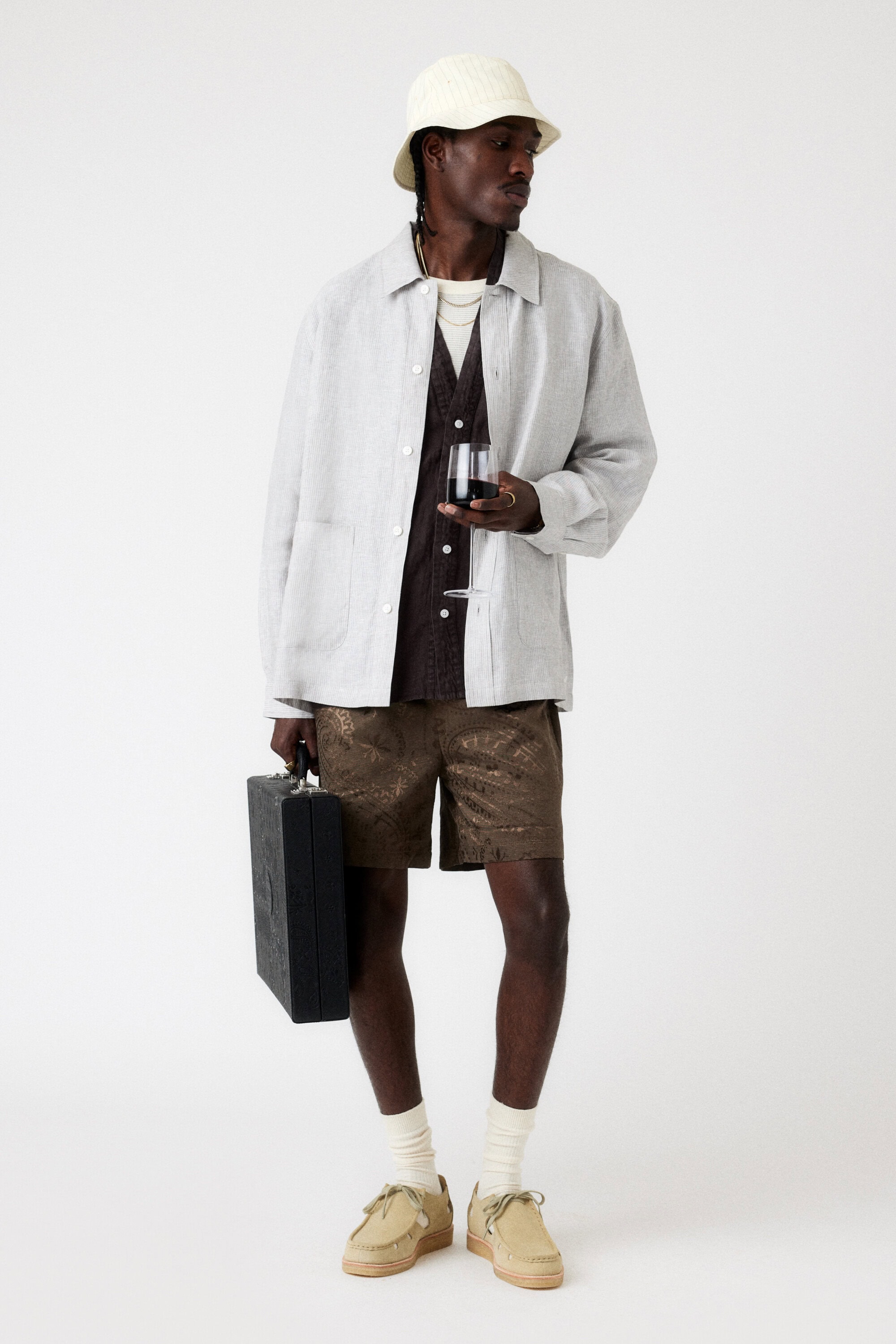 Full-shot of a Black man in a light jacket, patterned shorts, bucket hat, and loafers holding a briefcase and a glass of red wine in a studio.