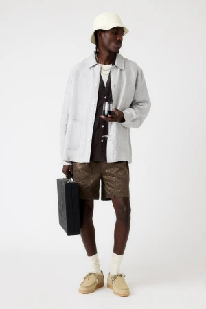 Full-shot of a Black man in a light jacket, patterned shorts, bucket hat, and loafers holding a briefcase and a glass of red wine in a studio.