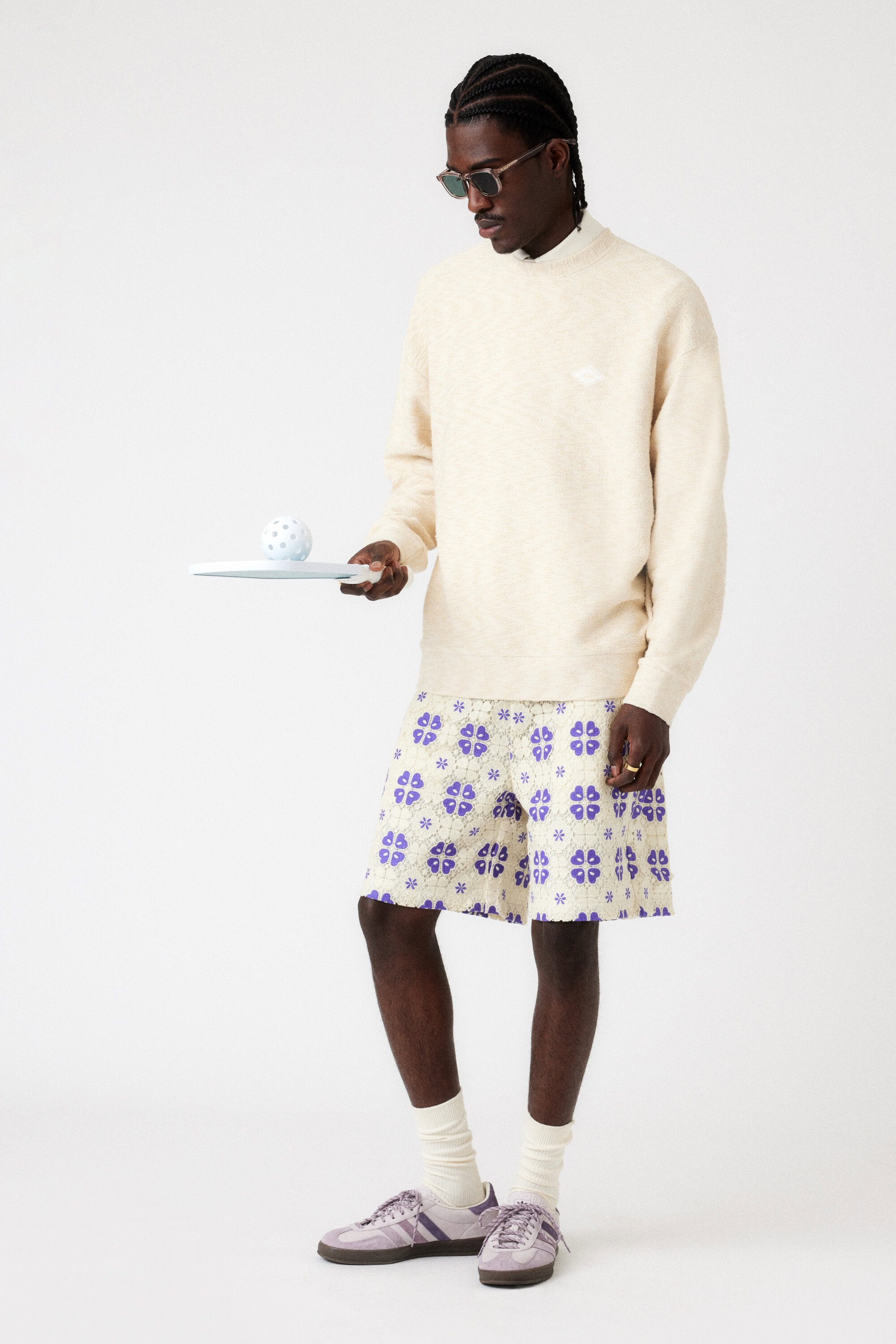 Man in light sweater and patterned shorts holds plate with ball in a studio setting with neutral colors, accessorized with sunglasses.