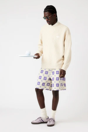 Man in light sweater and patterned shorts holds plate with ball in a studio setting with neutral colors, accessorized with sunglasses.