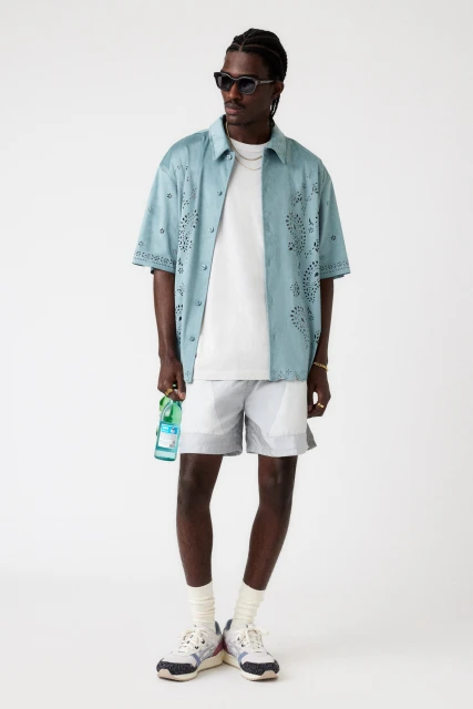 Full shot of a dark-skinned male model in an open aqua shirt, light shorts, and athletic shoes, holding a glass bottle against a white background.