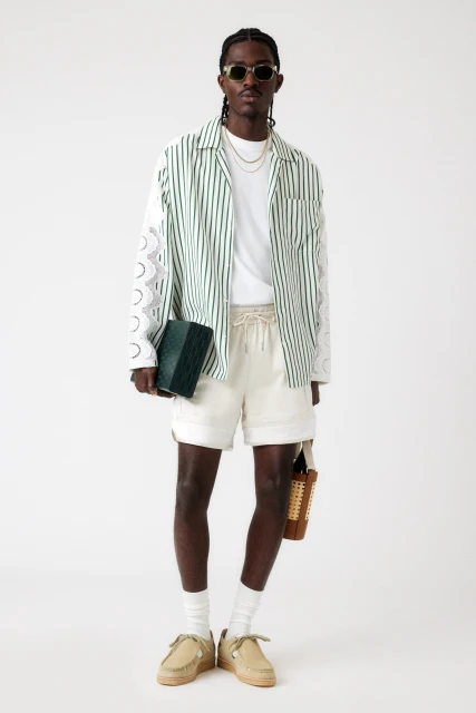 An adult Black man in a patterned shirt, shorts, and loafers stands in a full shot in a bright studio, holding two tote bags.