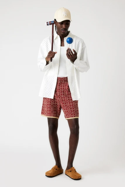 An adult Black male poses in a photo studio wearing an open white eyelet shirt, printed shorts, tan clogs and a tan baseball cap.