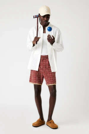 An adult Black male poses in a photo studio wearing an open white eyelet shirt, printed shorts, tan clogs and a tan baseball cap.