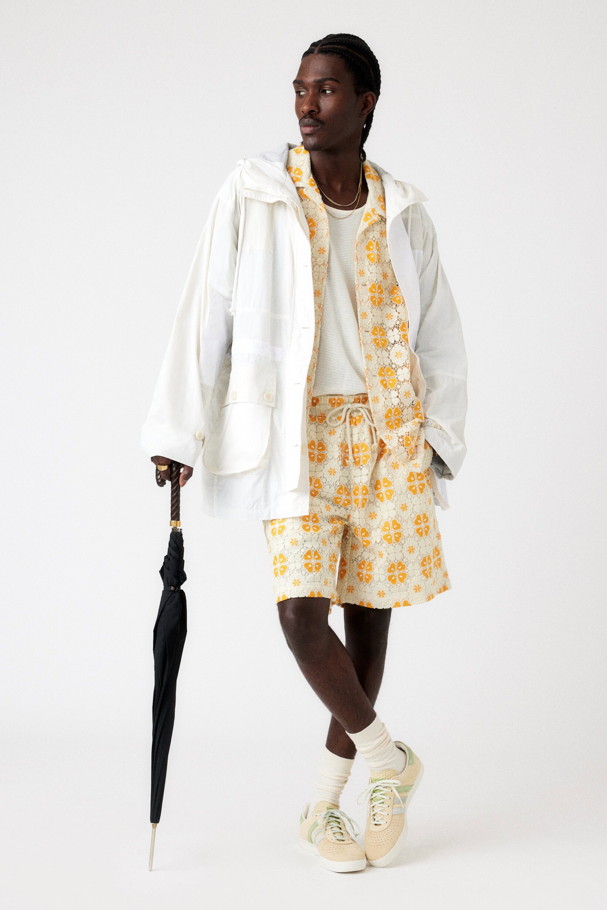 A black male poses in a white jacket, patterned shorts, and sneakers, holding a black umbrella against a clean, light background.
