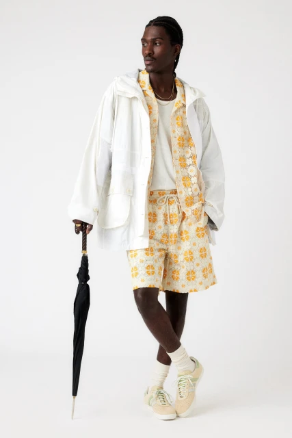 A black male poses in a white jacket, patterned shorts, and sneakers, holding a black umbrella against a clean, light background.